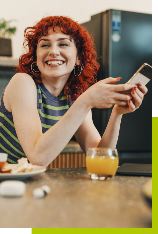Fröhliche Frau mit Smartphone am Küchentresen Junge Frau mit roten Locken sitzt lachend am Küchentresen, hält ein Smartphone in der Hand und ein Glas Orangensaft steht vor ihr.