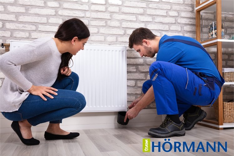 Heizkörper richtig entlüften Ein Mitarbeiter von Team Hörmann in blauer Arbeitskleidung entlüftet einen Heizkörper, neben ihm kniet eine junge Frau.