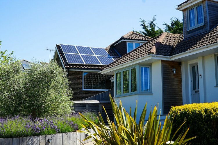 PV-Anlagen ohne Einspeisevergütung Ein Wohnhaus mit PV-Anlage auf dem Dach, im Vordergrund Gärten, im Hintergrund blauer Himmel.