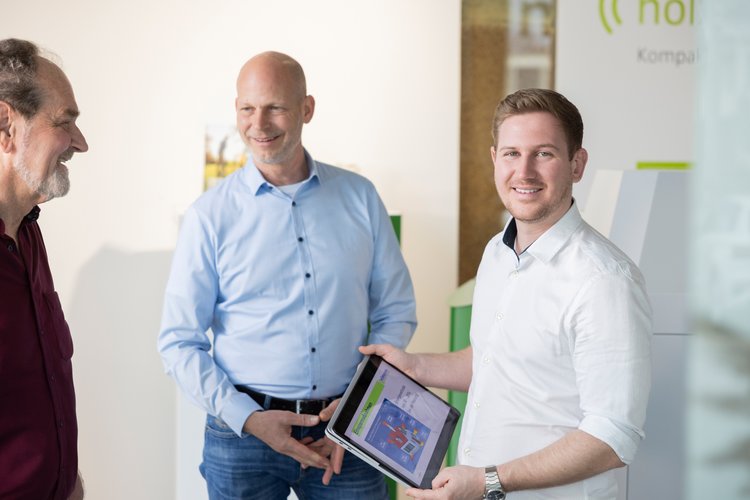 Lächelnde Kollegen bei Tablet-Präsentation im Büro Drei Männer stehen in einem hellen Büro, einer zeigt ein Tablet mit farbiger Darstellung und alle lachen freundlich.